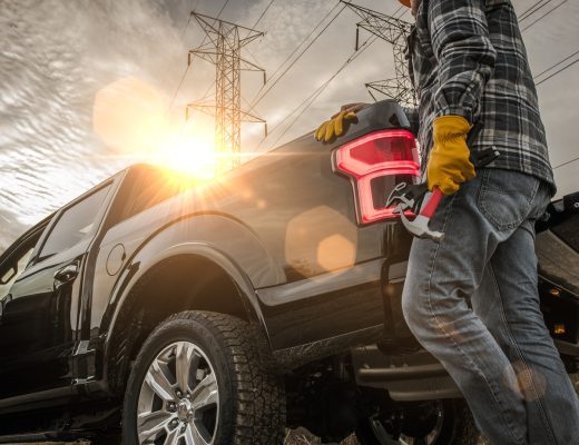 A man wearing yellow gloves and a helmet, holding a hammer while touching the back of a black pickup truck.