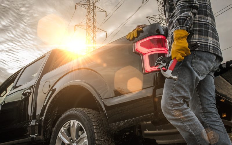 A man wearing yellow gloves and a helmet, holding a hammer while touching the back of a black pickup truck.