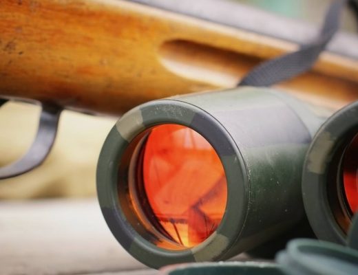 Binoculars, a shotgun, and hunting gear are placed together on top of a wooden table in an outdoor setting.