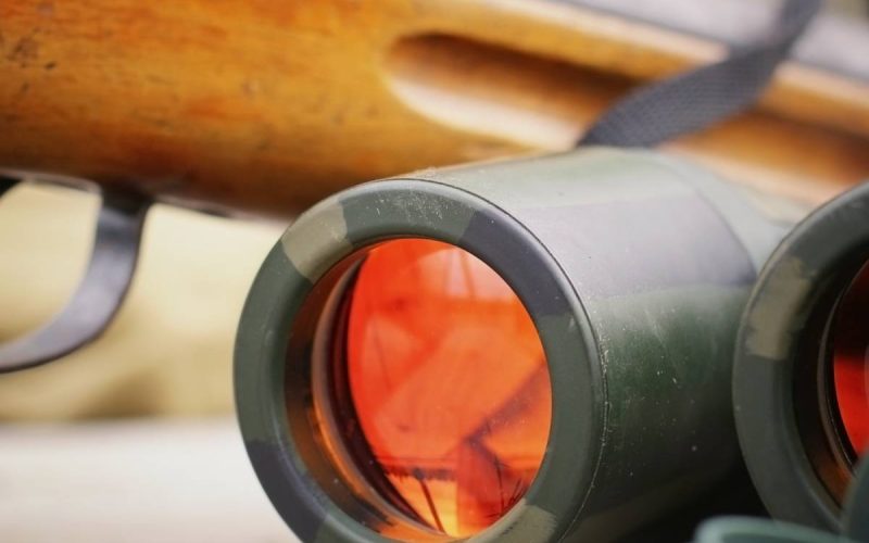 Binoculars, a shotgun, and hunting gear are placed together on top of a wooden table in an outdoor setting.