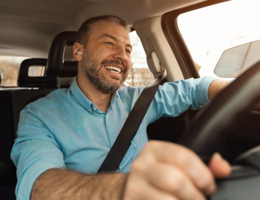 A smiling man sitting in the driver's seat of a vehicle with one hand on the wheel and the other resting on the window.