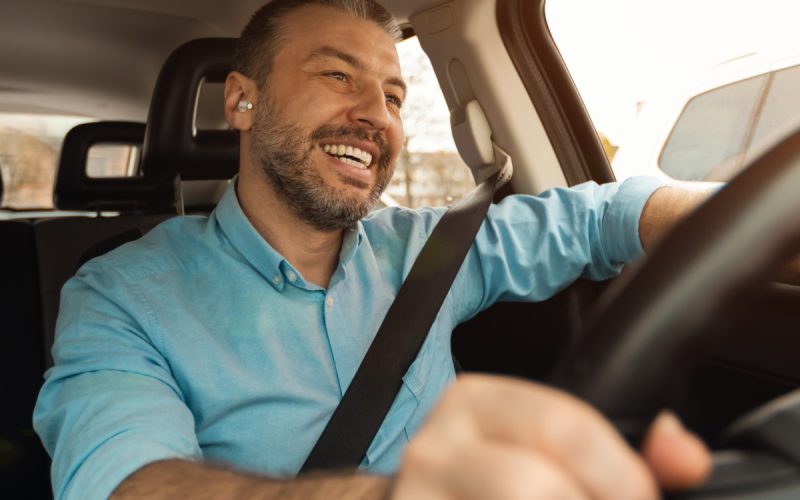 A smiling man sitting in the driver's seat of a vehicle with one hand on the wheel and the other resting on the window.
