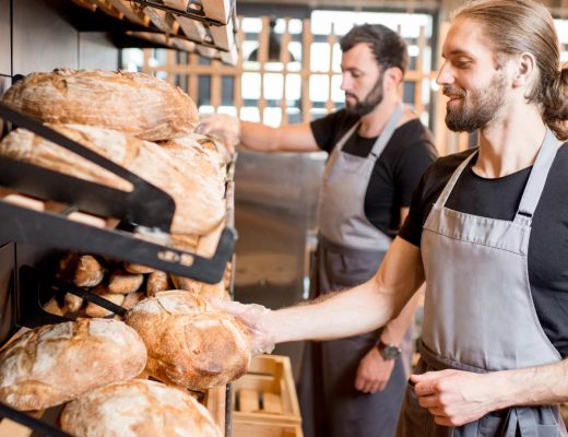Two men wearing black T-shirts and gray aprons are placing round loaves of bread into wooden crates at a bakery.