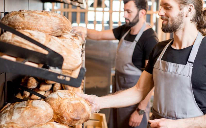 Two men wearing black T-shirts and gray aprons are placing round loaves of bread into wooden crates at a bakery.
