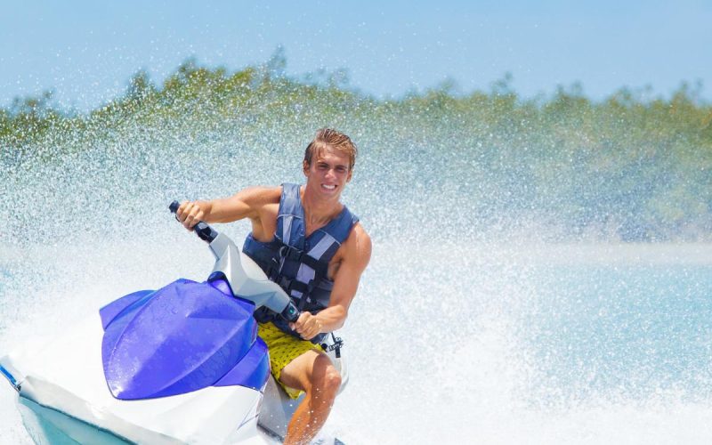 Man riding a jet ski on the water, wearing sunglasses and a life jacket, with small waves splashing around him.
