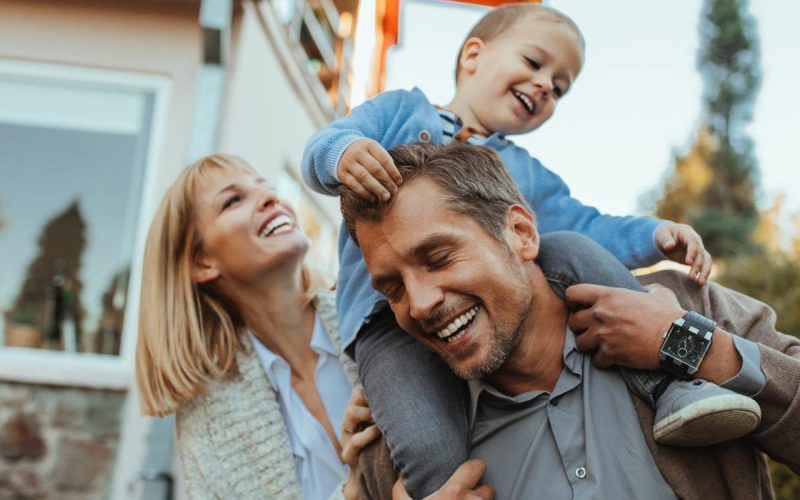 A man is carrying a child on his shoulders as a woman is walking next to him in an outdoor environment.