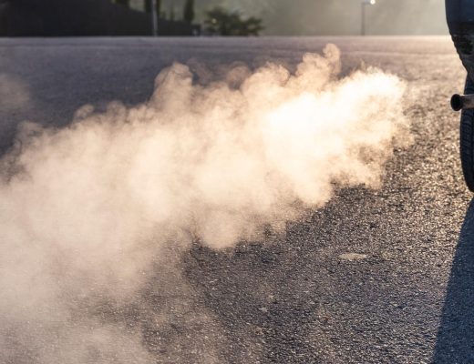 A cloud of white exhaust trailing behind the tailpipe of a truck in a sunlit parking lot in the morning.