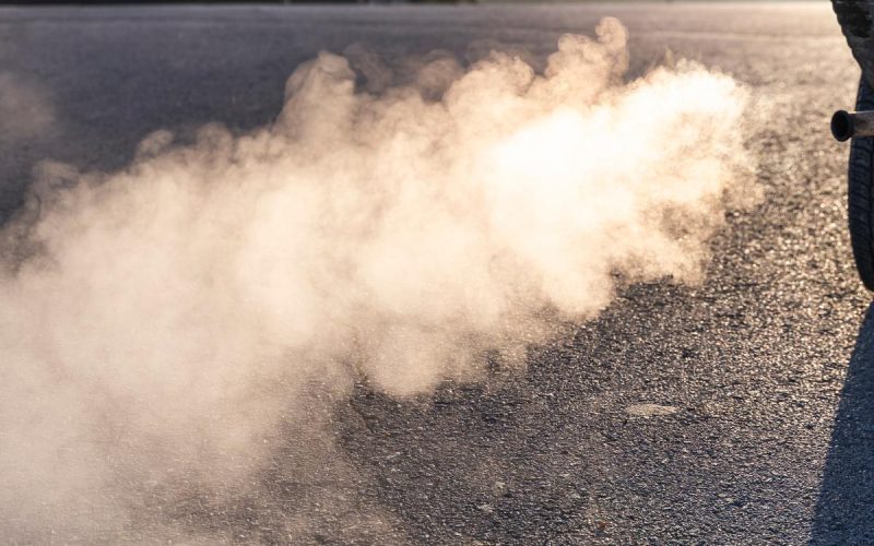 A cloud of white exhaust trailing behind the tailpipe of a truck in a sunlit parking lot in the morning.