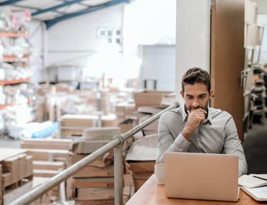 A man sits at a desk using a laptop in a warehouse office, with shelves, boxes, and inventory behind him.