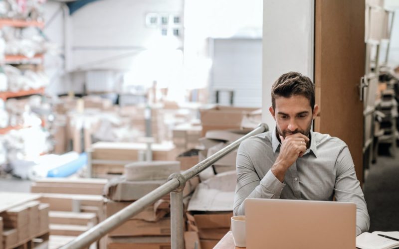 A man sits at a desk using a laptop in a warehouse office, with shelves, boxes, and inventory behind him.