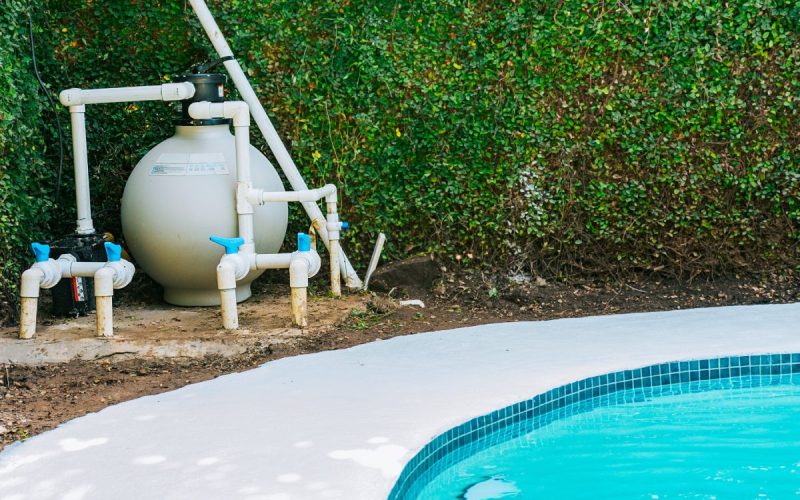 A pool equipment setup, including a filter, pump, and various pipes, on a concrete pad next to a swimming pool.