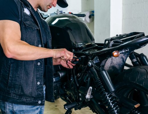 A man wearing a ballcap and a denim vest while using a hand tool to work on part of a black motorcycle.