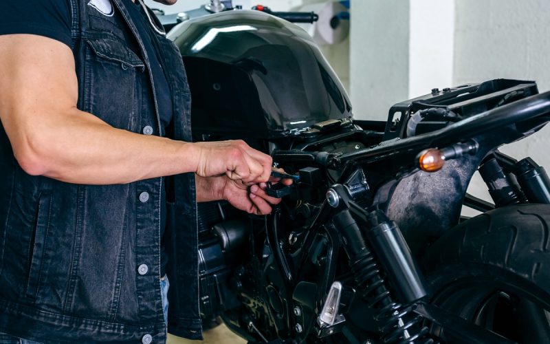A man wearing a ballcap and a denim vest while using a hand tool to work on part of a black motorcycle.