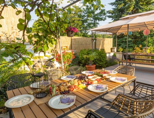An outdoor patio with a focus on a large, rectangular dining table with food and plates on it, surrounded by chairs.