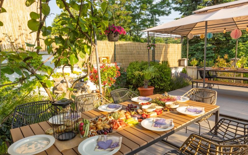 An outdoor patio with a focus on a large, rectangular dining table with food and plates on it, surrounded by chairs.