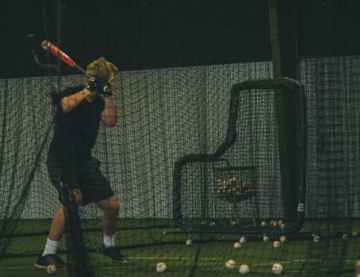 A young baseball player in a swing stance while standing in a batting cage with balls scattered on the floor.