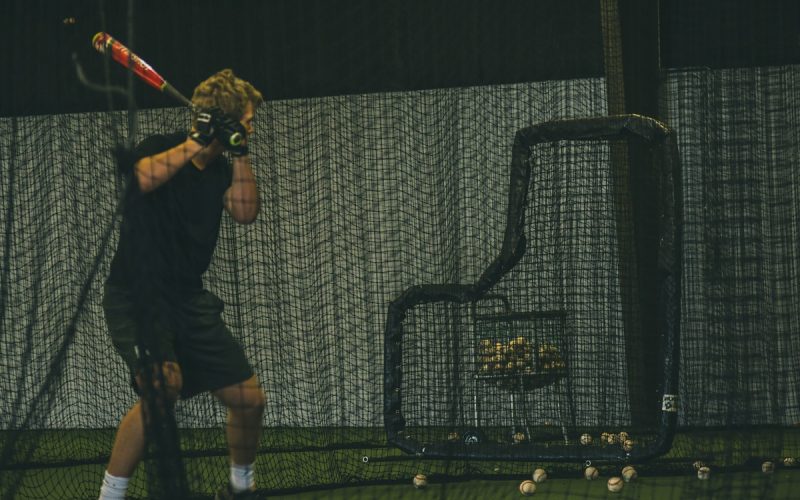 A young baseball player in a swing stance while standing in a batting cage with balls scattered on the floor.