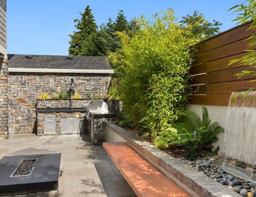 An enclosed backyard space with an outdoor kitchen, a fire feature with a bench seating area, and a waterfall feature.