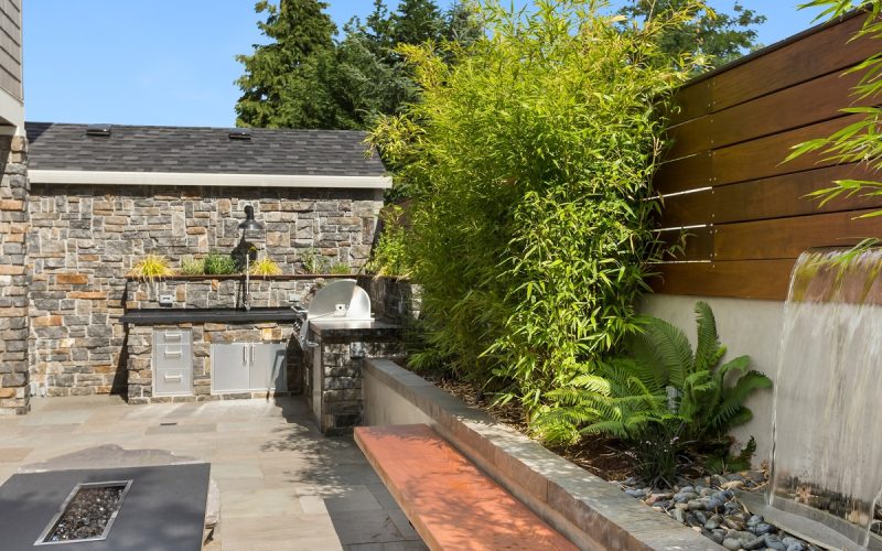 An enclosed backyard space with an outdoor kitchen, a fire feature with a bench seating area, and a waterfall feature.
