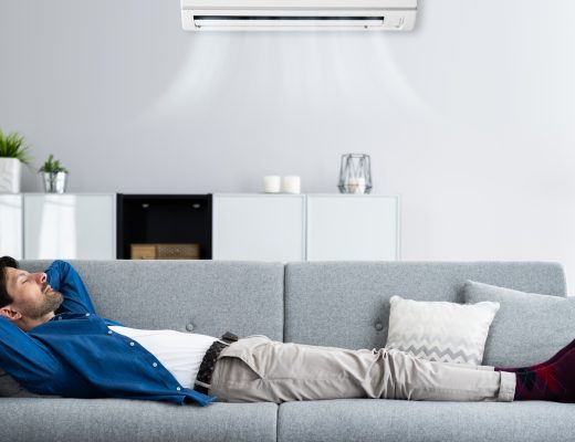 A man lies on a gray couch with his arms behind his head under a wall-mounted air conditioner in a living room.