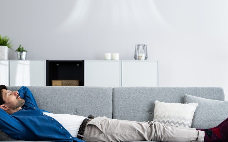 A man lies on a gray couch with his arms behind his head under a wall-mounted air conditioner in a living room.