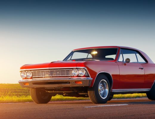 A classic red car parked on an asphalt road. A field of green grass shines in the sun behind the car.