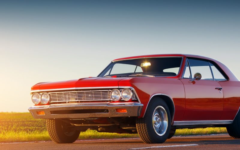A classic red car parked on an asphalt road. A field of green grass shines in the sun behind the car.