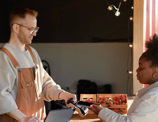 A man in an apron using a card reader as a woman holds a payment card at a wooden produce stall with strawberries.