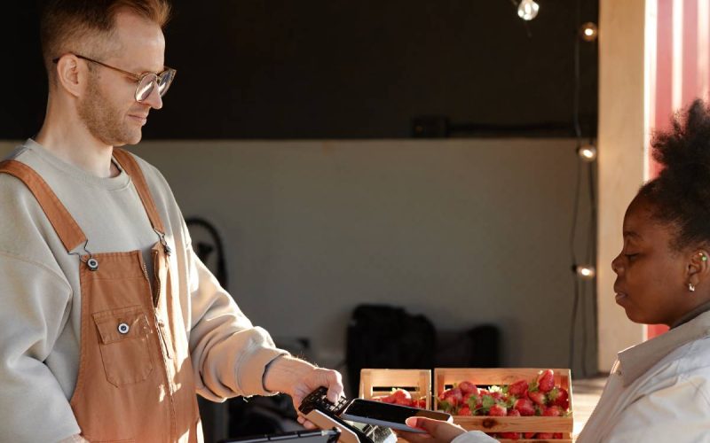A man in an apron using a card reader as a woman holds a payment card at a wooden produce stall with strawberries.