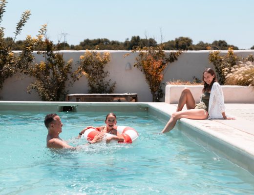 A woman sits on the edge of a pool surrounded by white concrete and a privacy wall. Two people are swimming in the water.