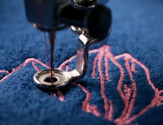 A close-up of a silver embroidery machine needle stitching a pink design on textured dark blue fabric.