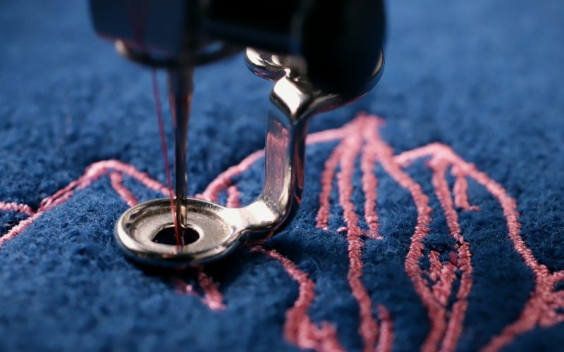A close-up of a silver embroidery machine needle stitching a pink design on textured dark blue fabric.
