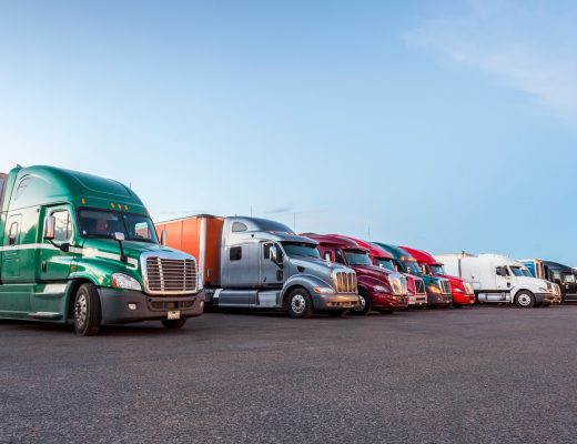 Several semi-trucks in different colors parked in a row on a large parking lot next to a white building.