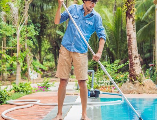 A man standing by the edge of an in-ground residential pool, holding a long pole attached to a hose in the water.