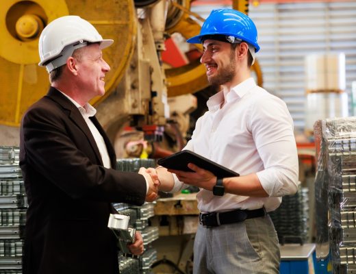 Two men in business attire and hard hats shaking hands in front of large equipment. One man is holding a tablet.