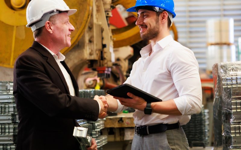 Two men in business attire and hard hats shaking hands in front of large equipment. One man is holding a tablet.