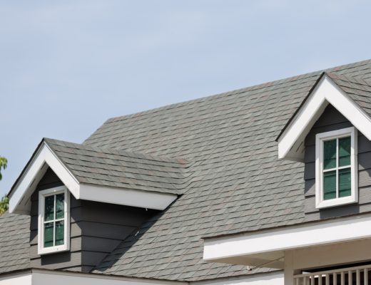 A gray asphalt shingle roof on a dark-sided home with dormer windows beneath a clear blue sky and leafy trees.