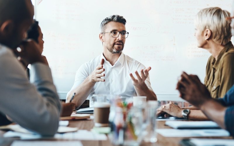 A man with glasses speaking with raised hands to people seated around a meeting table in an office with a whiteboard.