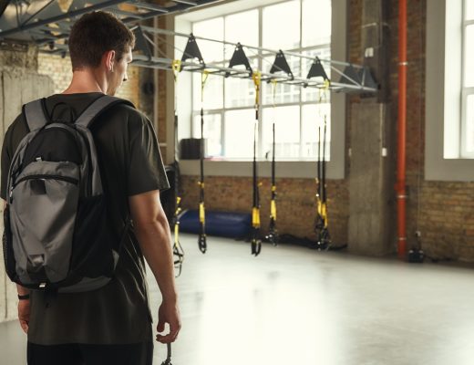 A man with a backpack stands in a modern gym space with suspension training straps and equipment in the background.