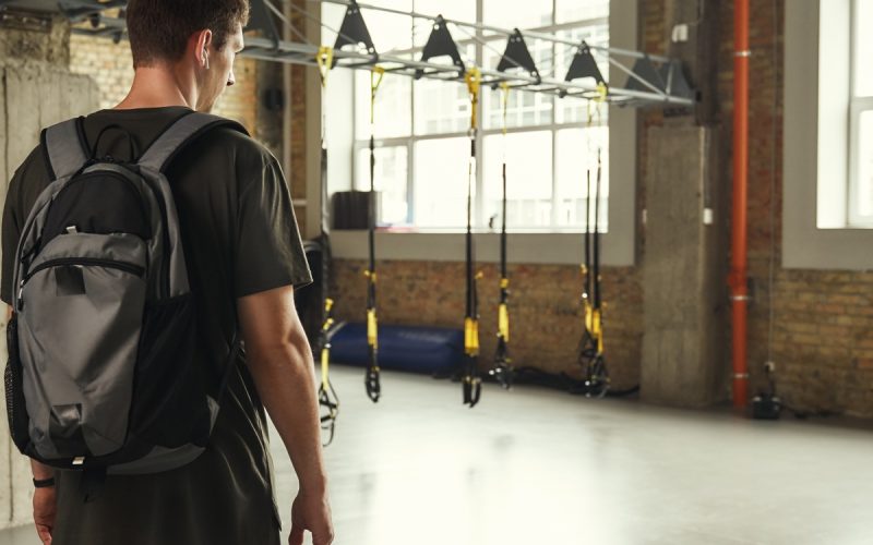 A man with a backpack stands in a modern gym space with suspension training straps and equipment in the background.