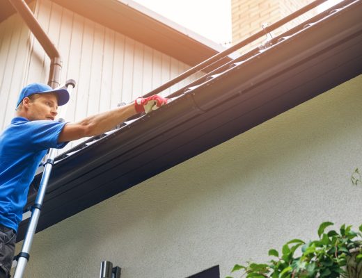 Worker on a ladder cleaning a home's roof gutter beside black downspouts, with a chimney and green shrubs in view.