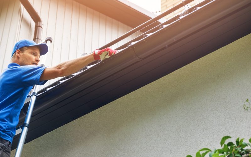 Worker on a ladder cleaning a home's roof gutter beside black downspouts, with a chimney and green shrubs in view.
