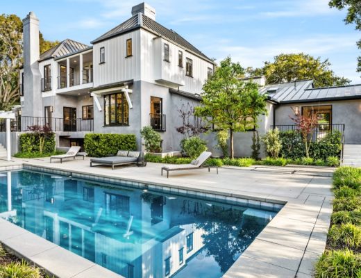 The backyard of a large white home with a swimming pool, pergola, landscaping, and lounge chairs along one side of the pool.