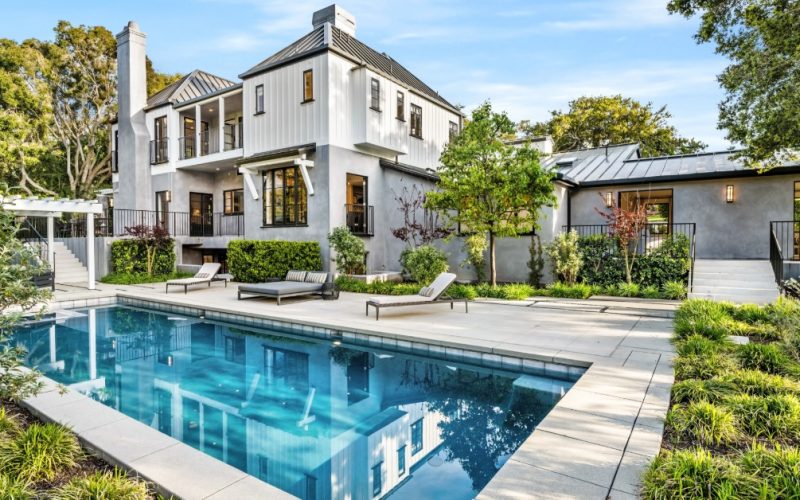 The backyard of a large white home with a swimming pool, pergola, landscaping, and lounge chairs along one side of the pool.
