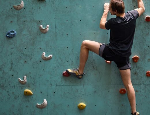 A person wearing dark clothing climbs a teal indoor rock wall with colorful handholds and one foot stretched down.