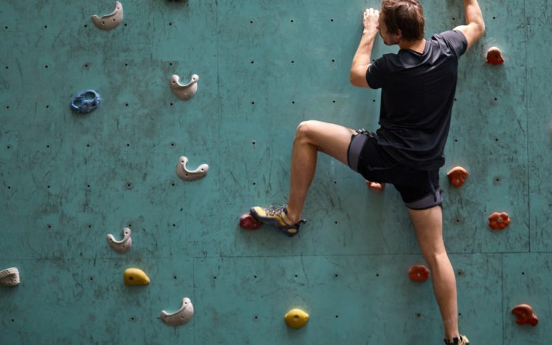 A person wearing dark clothing climbs a teal indoor rock wall with colorful handholds and one foot stretched down.