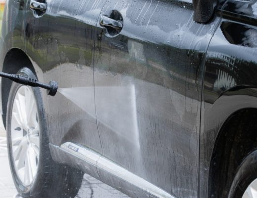 A close-up of a pressure washer spraying water onto the side of a black SUV parked in a driveway during an exterior car wash.