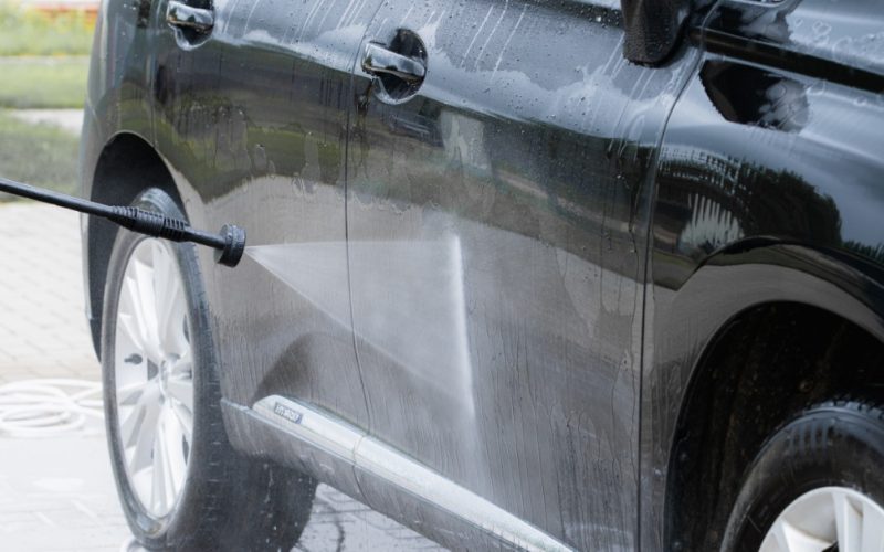 A close-up of a pressure washer spraying water onto the side of a black SUV parked in a driveway during an exterior car wash.