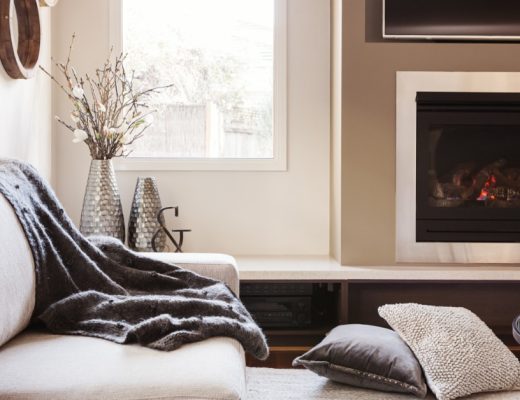 A comfy living room with a white sofa and coffee table. A fireplace and window line the wall of the room.