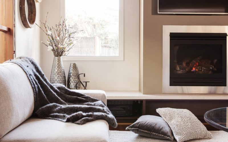 A comfy living room with a white sofa and coffee table. A fireplace and window line the wall of the room.